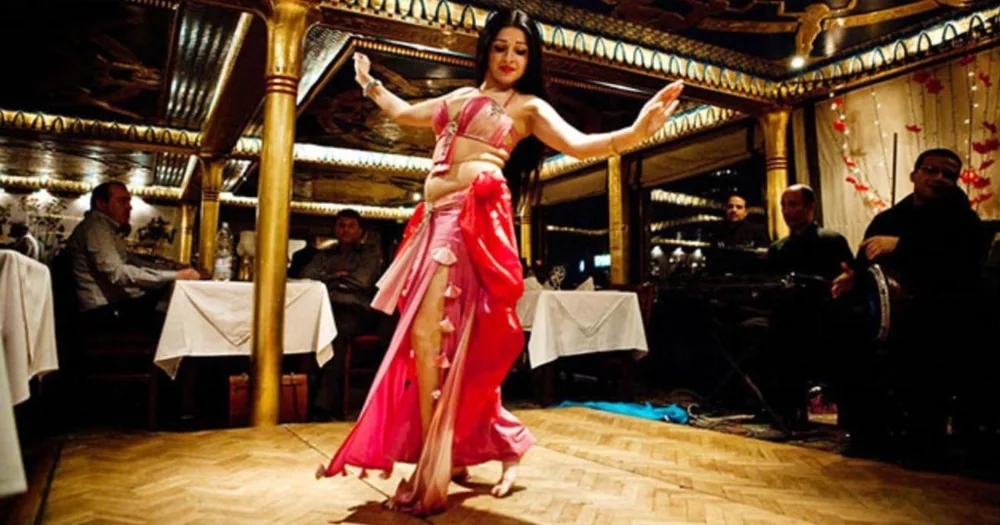 A belly dancer in a pink costume performing between tables on a Nile dinner boat, musicians behind
